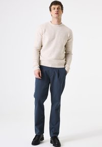 Beige gebreide trui met een ronde halslijn, gecombineerd met donkerblauwe geplooide broeken en zwarte veterschoenen. Eenvoudige en casual outfit.