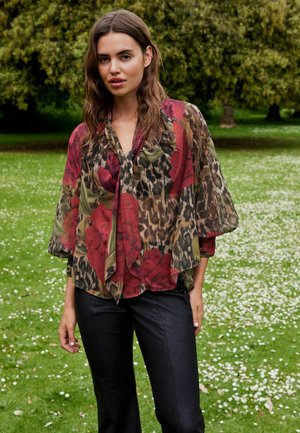 Top floreale trasparente con vivaci rose rosse e stampa animale, con colletto a lacci e maniche ampie. Indossato su pantaloni scuri e aderenti.