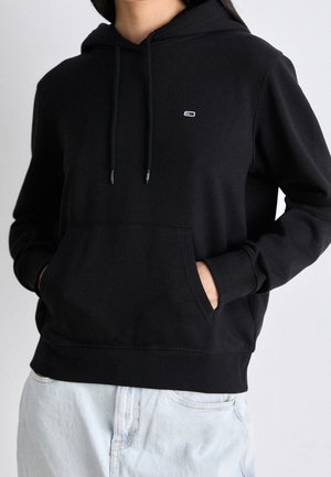 Persona che indossa una felpa nera con cappuccio, tasca a marsupio e piccolo logo sul petto, abbinata a jeans azzurri, con le mani nelle tasche.