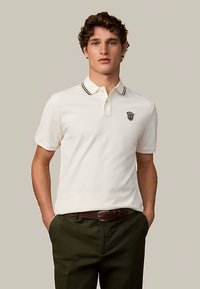 Polo bianca con bordino nero sul colletto e logo sul petto. Abbinata a pantaloni verde scuro e cintura marrone.