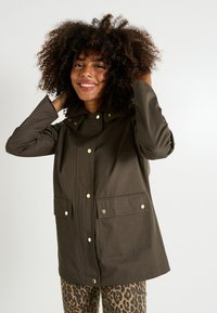 Chaqueta impermeable de color verde oliva con un cuello alto, dos bolsillos frontales y botones de presión dorados. La lleva puesta sobre unos pantalones de estampado de leopardo.