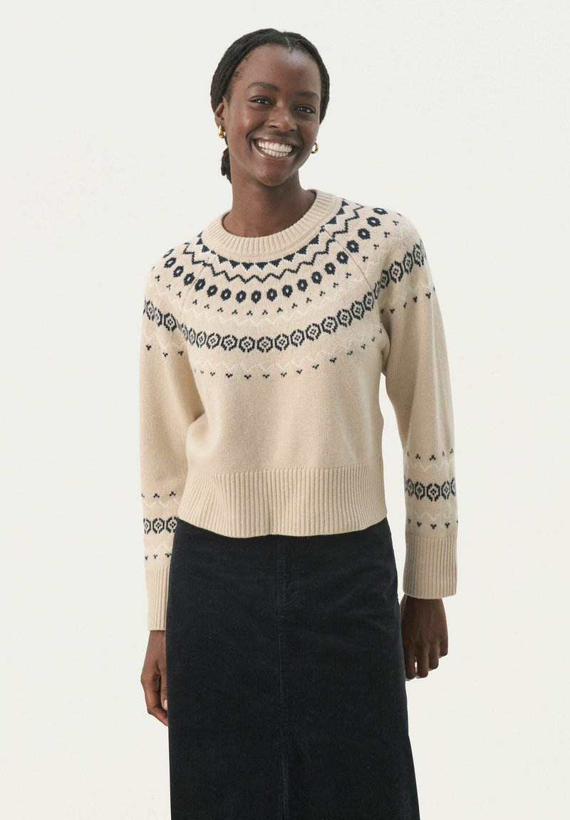 Pull beige à col rond, ornements marins à motifs autour des épaules et des manches, ourlet côtelé, texture douce, associé à une jupe noire.