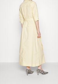 Person i en ljusgul lång klänning med trekvartslånga ärmar och zebramönstrade kitten heel-skor, stående framför en vit bakgrund.