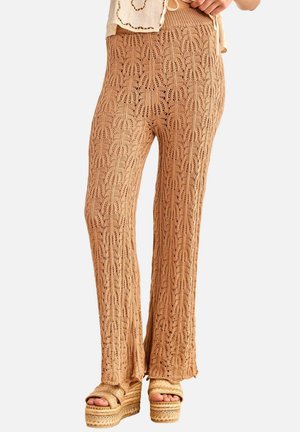 Mujer con pantalones beige de punto de pierna ancha y sandalias beige de plataforma con textura tejida, de pie sobre un fondo blanco.
