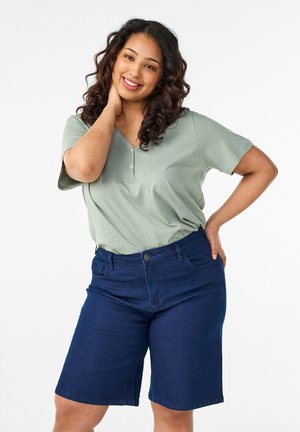 Smilende kvinde med krøllet hår iført lysegrøn kortærmet skjorte og mørkeblå knælange denimshorts, poserer med hånden på nakken og hoften.