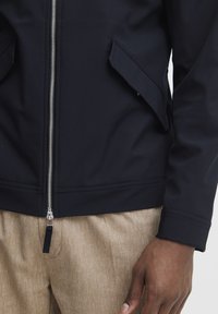 Giacca navy con zip frontale, due tasche a battente inclinate e un design aderente. Indossata sopra pantaloni beige strutturati.