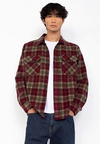 Weinrote Flanellhemd im Karo-Muster mit grünen und cremefarbenen Akzenten, zwei Fronttaschen und schwarzen Knöpfen. Über einem weißen T-Shirt getragen.