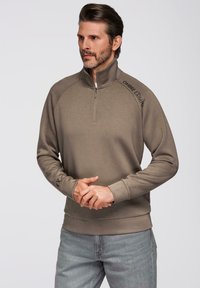 Sudadera de color caqui con cremallera de un cuarto, mangas raglán, puños y dobladillo acanalados, con detalles de logotipo en el cuello y la manga. Textura suave.