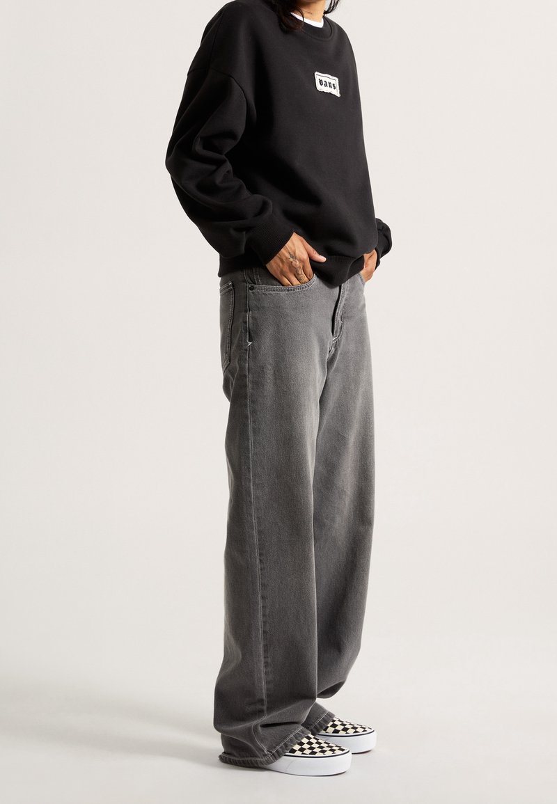 Personne portant un sweat-shirt noir avec un logo, un jean gris ample et des chaussures slip-on à motif damier noir et blanc, debout les mains dans les poches.