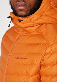 Orange isoleret jakke med hætte, med quiltet design, en frontlynlås og "PeakPerformance" logo broderet på brystet.