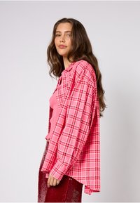 Różowa koszula w kratę z długimi rękawami i kołnierzem, nałożona na różowy top na ramiączkach. Gładka tkanina i luźny krój, a poniżej czerwona cekinowa spódnica.