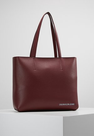 Borsa tote in pelle testurizzata bordeaux con due spallacci, forma rettangolare e piccolo logo bianco "Calvin Klein Jeans" nell'angolo inferiore.