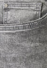 Primer plano de unos jeans de mezclilla gris, que presentan un bolsillo delantero, costuras visibles y un ligero desvanecimiento para aportar textura. Se incluye detalle del botón superior plateado.