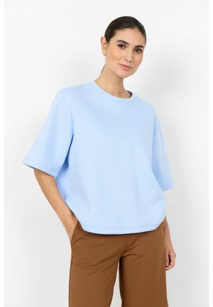 Woman wearing a light blue oversized short-sleeve top and brown pants, posing with one hand in pocket against a white background.