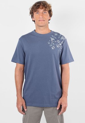 Homme aux cheveux bouclés portant un t-shirt bleu avec un petit imprimé floral sur l'épaule droite, debout devant un fond clair uni.