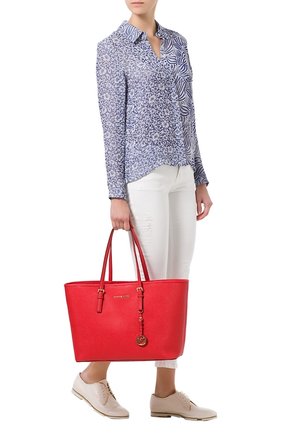 Femme en pantalon blanc et chemise bleue à motifs, tenant un grand sac fourre-tout rouge et portant des chaussures beiges, debout devant un fond blanc.