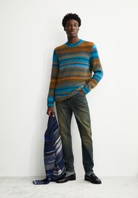 Strickpullover mit horizontalen Streifen in Blau, Orange und Braun, kombiniert mit grünen Jeans und schwarzen Loafers, hält einen blau und grau gestreiften Schal.