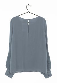 Blusa grigia a maniche lunghe con scollo a barca, dettagli in pizzo testurizzato sulle maniche e chiusure a bottone sui polsini. Presenta un dettaglio con bottone sul retro.