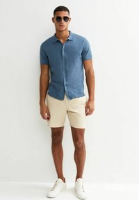 Camisa de manga corta, abotonada, en tela azul claro, combinada con pantalones cortos beige y zapatillas blancas. Gafas de sol negras y redondas completan el conjunto.