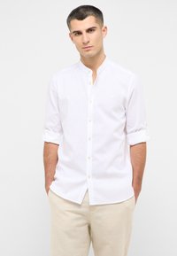 Camicia bianca a maniche lunghe con colletto alla cinese, chiusura a bottoni e maniche arrotolate. Il tessuto ha una texture liscia e leggera.