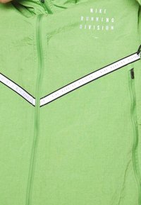 Veste verte avec une finition texturée, dotée d'une fermeture éclair à l'avant, d'une bande de marque noire et blanche traversant la poitrine, et d'une capuche.