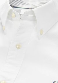 Weißes Baumwollhemd mit Button-Down-Kragen, ausgestattet mit Perlmuttknöpfen und einem gestreiften Futter am Kragen. Stickereidetail mit Logo auf der Brust.