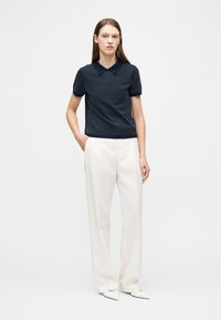 Γυναίκα που στέκεται φορώντας μπλε σκούρο πόλο με κοντά μανίκια και λευκά πουά, φαρδιά λευκά παντελόνια και λευκά μυτερά παπούτσια, μπροστά σε απλό λευκό φόντο.