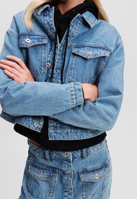 Ljusblå denimjacka med en kort design, tryckknappar och två bröstfickor, lagd över en svart huva-svetetröja.