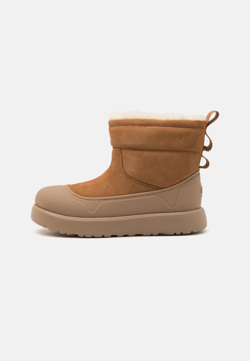 Bottine en daim marron avec une doublure en fausse fourrure crème, une semelle en caoutchouc beige et des boucles décoratives à l'arrière pour un enfilage facile.