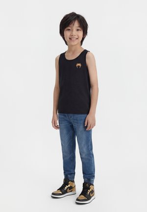 Garçon debout, portant un débardeur noir avec un petit logo orange, un jean bleu et des baskets montantes noires et dorées.