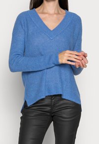 Blau gestrickter Pullover mit V-Ausschnitt, langen Ärmeln und seitlichen Schlitzen, aus weichem, strukturiertem Material. Getragen mit schwarzen Lederhosen.