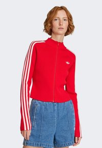 Rote, gerippte Zip-Jacke mit hohem Kragen und weißen Streifen an den Ärmeln. Kombiniert mit blauen Denim-Shorts.