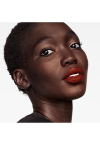 Gros plan sur une personne à la peau foncée avec des cheveux courts, présentant des lèvres rouges brillantes et un maquillage des yeux subtil en doré, sur fond blanc.