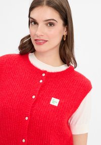 Eine Frau trägt eine leuchtend rote Strickweste mit Knöpfen über einem weißen Kurzarmoberteil und goldenen Creolen, lächelt leicht.