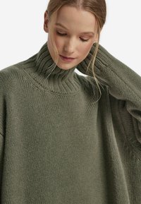 Oliwkowy sweter z golfem, dzianinowa tekstura, prążkowany kołnierz i mankiety, luźny krój, z widocznymi detalami szwów na ramionach.