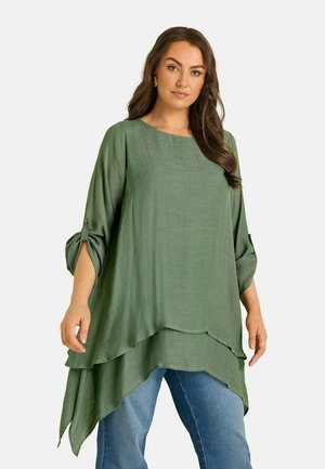 Donna che indossa una tunica larga a strati color verde oliva con maniche risvoltate ai 3/4 e jeans blu su uno sfondo chiaro e semplice.
