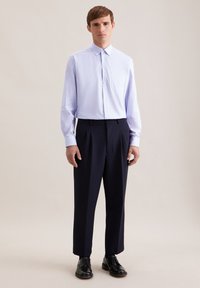 Camicia celeste con bottoni e taschino, abbinata a pantaloni plissettati blu navy scuro e scarpe di pelle nere. Design e colori semplici e puliti.