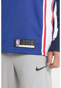 Primo piano di una persona che indossa una maglia NBA swingman blu, bianca e rossa e pantaloni grigio chiaro con il logo Nike nero.