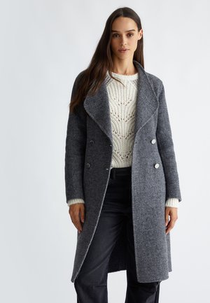 Cappotto grigio in misto lana con ampio bavero, design doppiopetto e due bottoni argentati. Indossato sopra un maglione lavorato a maglia color crema.