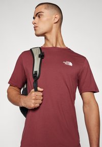 Burgunderfarget kortermet t-skjorte med rund hals og hvit logo. Modellen holder en svart bag med grå stropp og spenne.