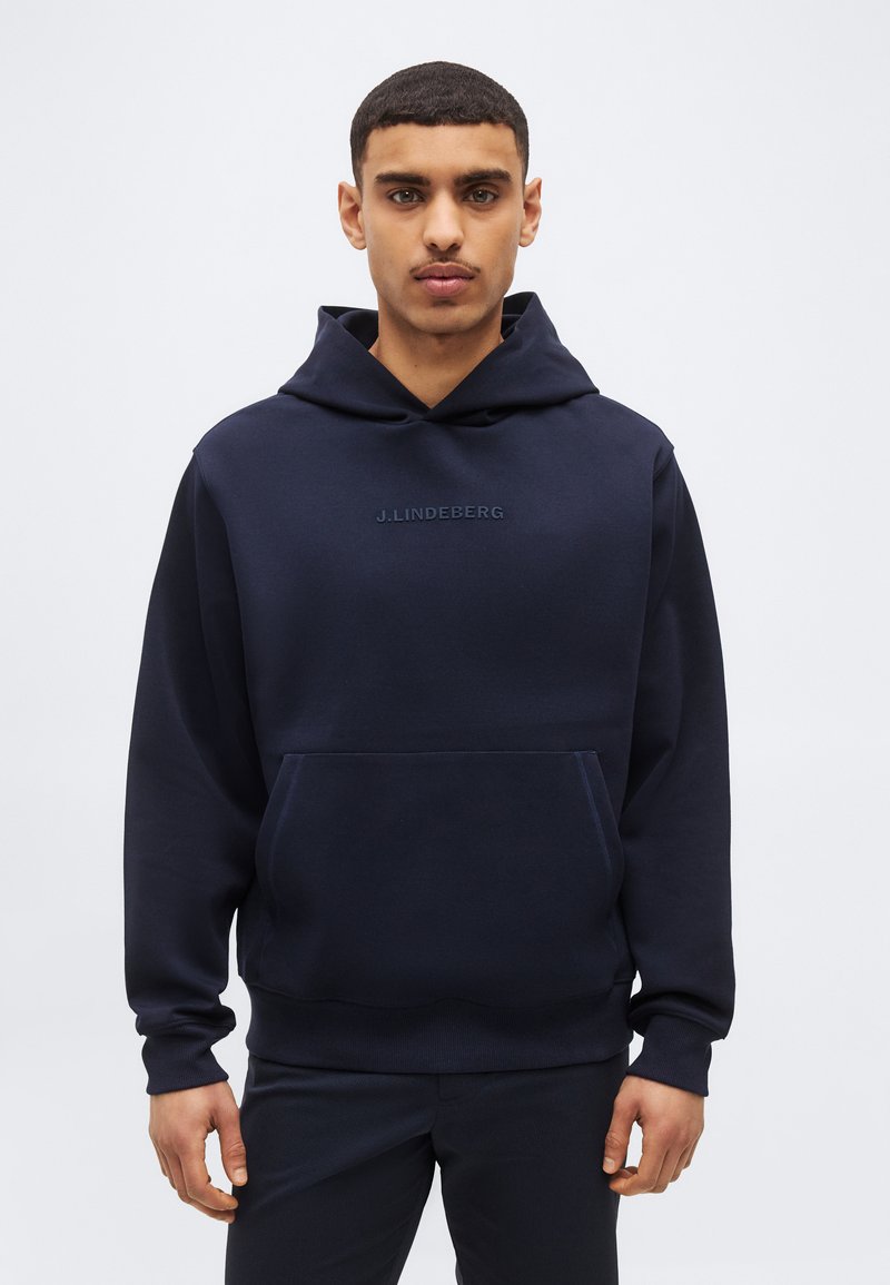 Νεαρός άνδρας που φοράει μπλε σκούρο hoodie J.Lindeberg με τσέπη καγκουρό, στέκεται μπροστά σε ένα απλό ανοιχτό γκρίζο φόντο.
