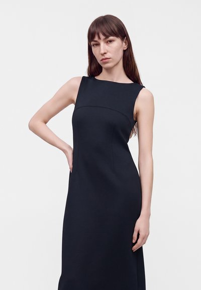 Femme aux longs cheveux bruns portant une robe noire sans manches, debout avec une main sur la hanche devant un fond uni et clair.