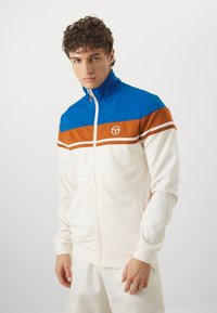 Chaqueta de chándal con cremallera en color crema con franjas horizontales azules y marrones. Presenta un cuello alto, bolsillos laterales y una tela texturizada.