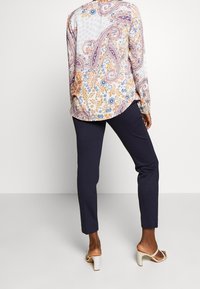 Blouse à manches longues, à motifs dans des tons clairs avec des dessins cachemire et floraux, associée à un pantalon bleu marine et des sandales blanches à talons blocs.