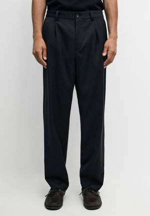 Mann trägt lockere schwarze plissierte Hose und dunkelbraune Schnürschuhe und steht vor einem schlichten Hintergrund.