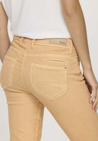 Jean en denim pêche clair au design ajusté, comportant deux poches arrière et une étiquette sur la ceinture. Texture lisse avec un minimum de détails.