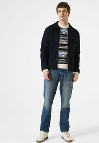 Man in een marineblauwe jas over een gebreid patroontrui, blauwe spijkerbroek en witte sneakers, staande tegen een effen witte achtergrond.