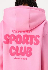 Rosa hoodie gjord av mjukt tyg med en stor, iögonfallande tryck "SPORTS CLUB", samt mindre text som innehåller "ellesse" och "sedan 1959."