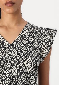 Svart-hvitt mønstrete topp med V-hals, volangermet armer og diamantformede motiver. Mykt stoff med teksturert overflate.