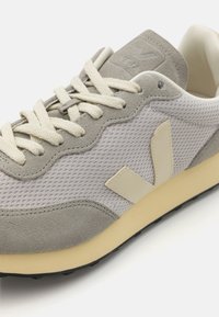 Sapatilhas cinzas com parte superior em malha e detalhes em camurça, atacadores em creme e uma sola de borracha macia. Logótipo em relevo na lingueta e na lateral.